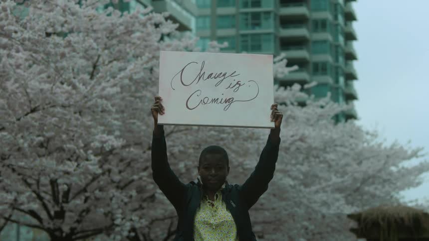 A black female protestor with yellow shirt and black leather jacket holds up banner saying change is coming