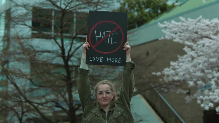 Young woman standing in front of a building lifting a protest sign against hate
