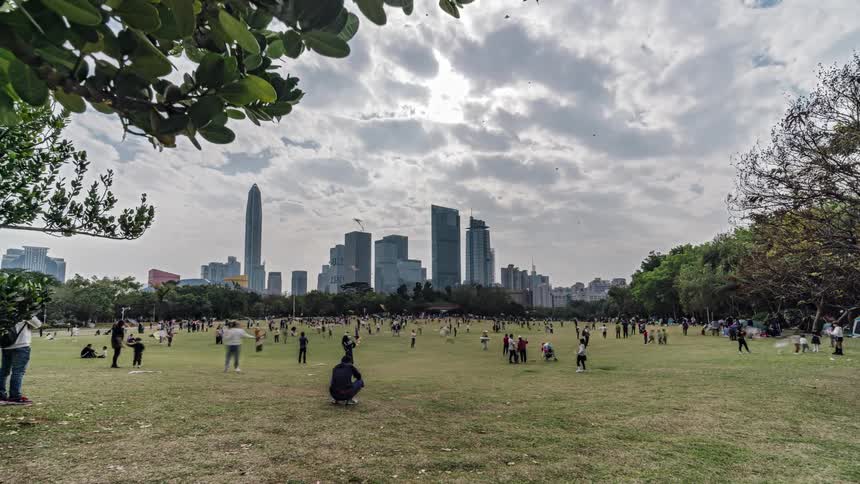 A large group of people fly kites in a large city park on a cloudy day