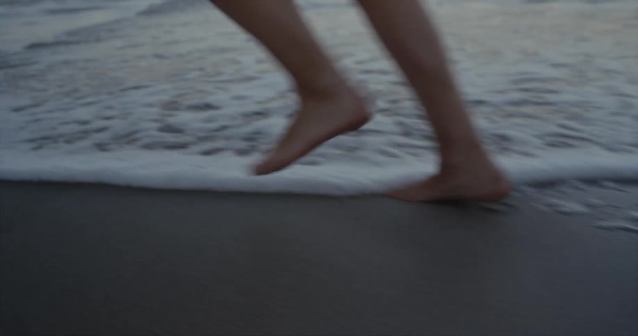 A person with bare feet is walking in the sand on a beach as the waves roll in