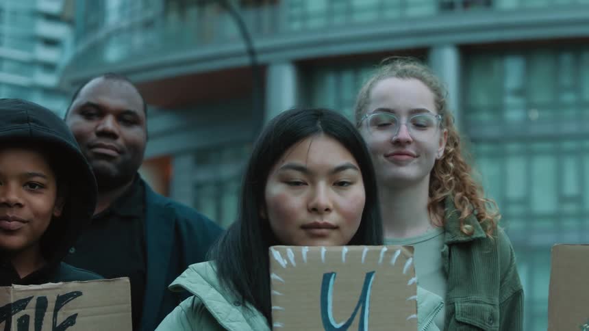 A group of activists hold signs while looking into the camera as they stand outdoors during the day