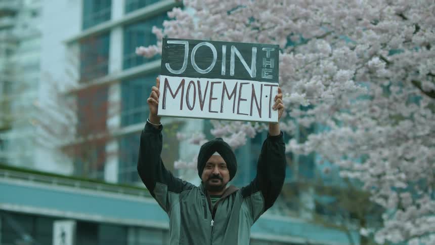 A smiling man raises a protest sign as he stands in front of a building