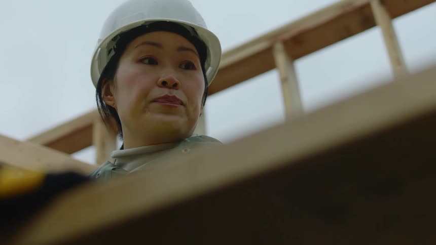 An asian woman wearing a white hardhat is speaking at a construction site