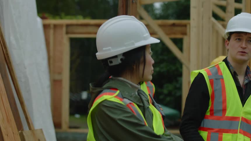 A construction crew is on a construction site and a male worker is speaking to an asian woman wearing a white hardhat