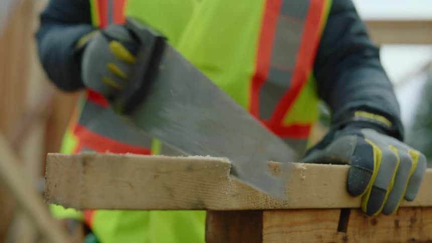 A construction worker cuts a wooden plank with a hand saw on a construction site during the day