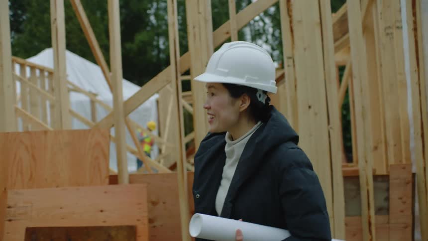 A construction worker shakes hands with an asian woman wearing a coat and a hardhat and blueprints rolled up under her arm