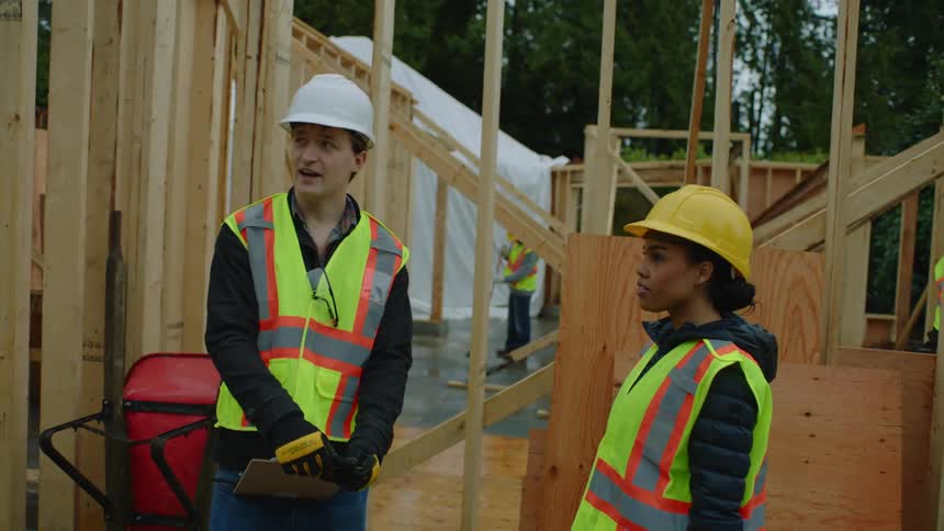 A female construction worker shakes hands with a male coworker carrying a clipboard and they stop to talk