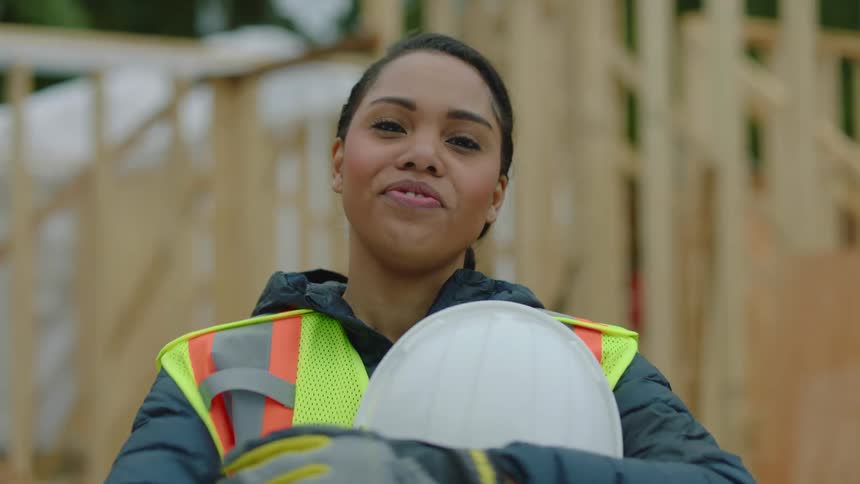 Young female construction worker standing on a construction site wearing a vest and taking out her safety helmet
