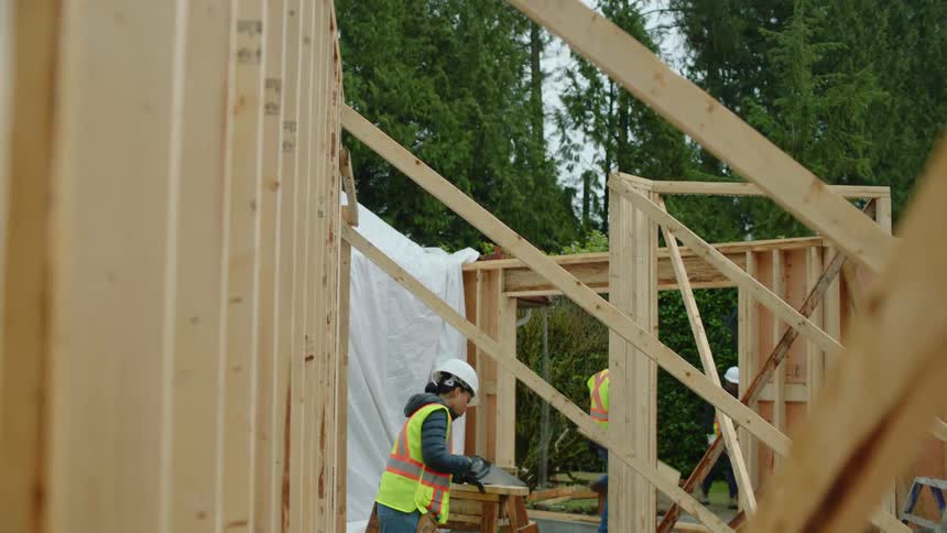 A female construction worker is sawing wood as other crew members work around the site