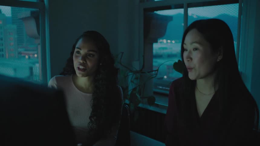 Two women holding a conversation while sitting at the desk in front of the computers next to two big windows
