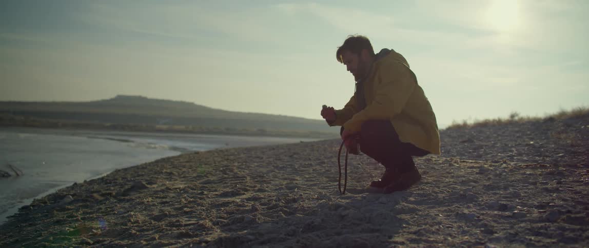 A man bends down and picks up a rock to skip across a lake