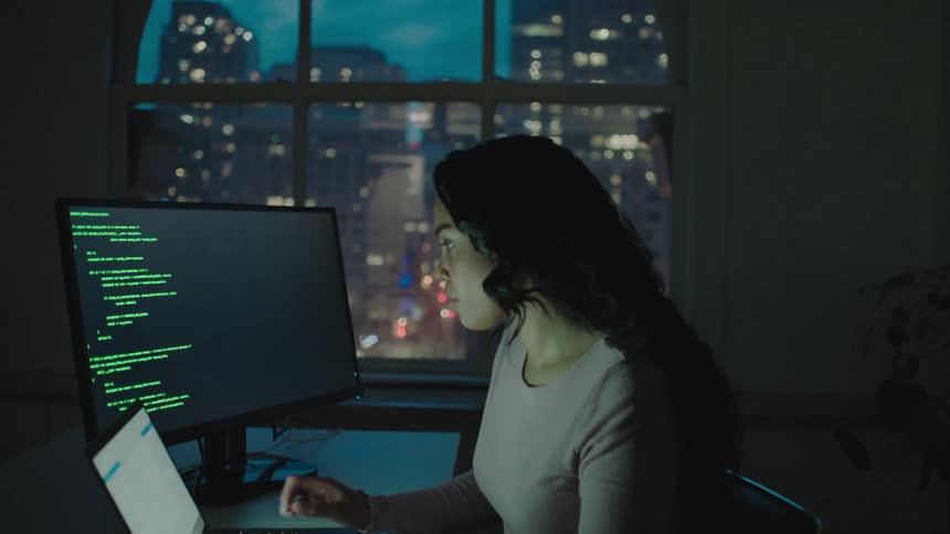 A woman sits at a desk with a laptop and a large computer monitor