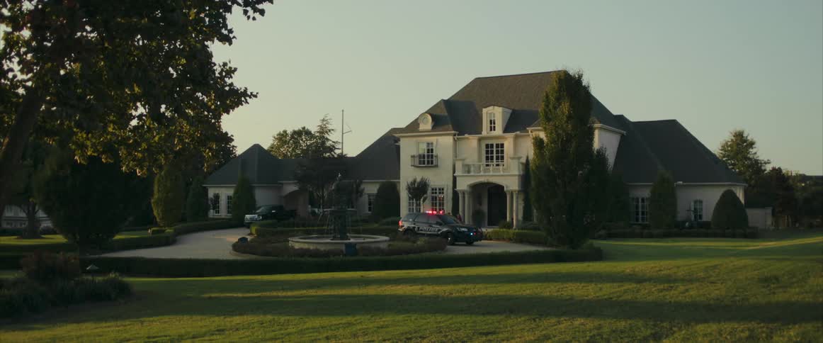 A cop leads a teenager out of a house toward a cop car as his mother follows behind him