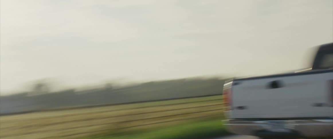 Teenagers driving a white pickup truck speeds by the camera and drives away down a country road