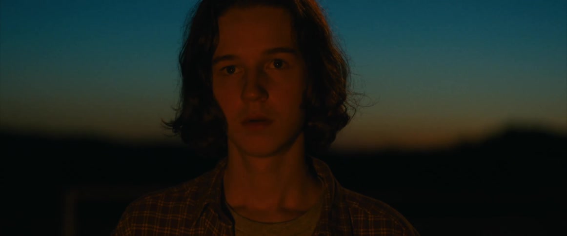 A pensive teenage boy staring at a fire outdoors during blue hour