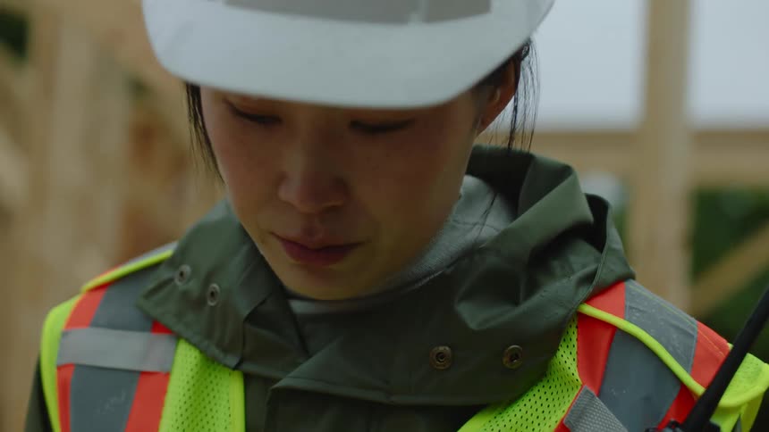 A female construction worker wearing a white hardhat is measuring and marking a piece of wood