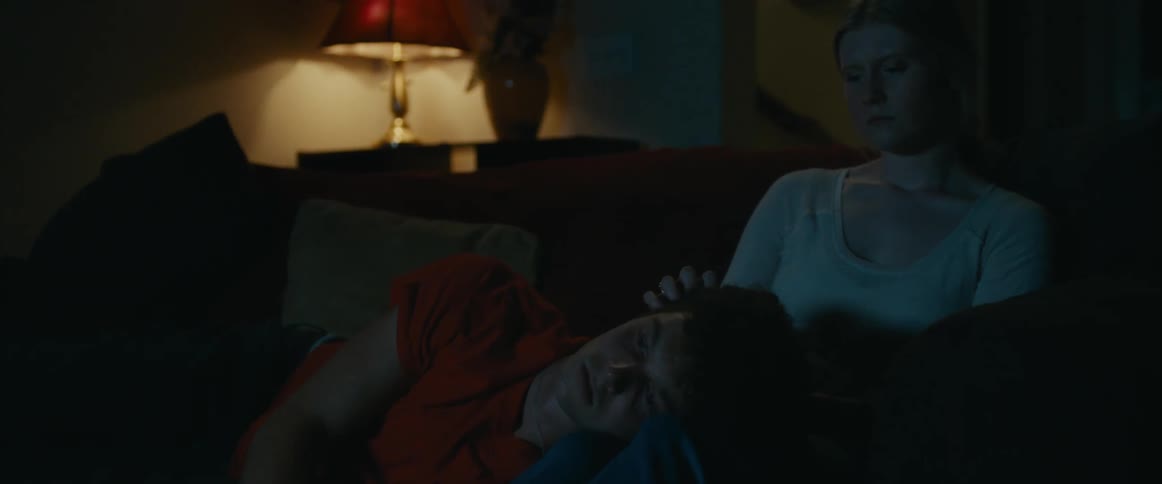 A woman sits on a couch at home at night and strokes a mans hair as he lies in her lap