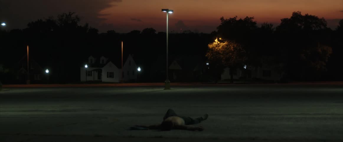 A shirtless man lying on his back in a parking lot at sunset