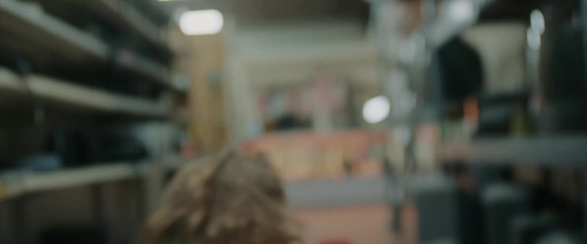 A teenage boy working in a store throws something off a shelf and pushes a car down an aisle and sits down and breathes deeply