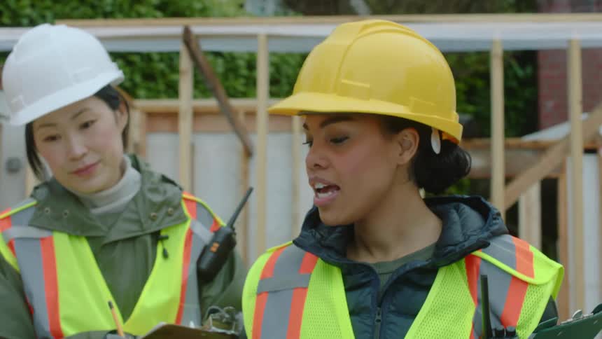 A female construction worker is carrying a notebook and walking around the construction site as she calls to an asian woman and speaks to the crew