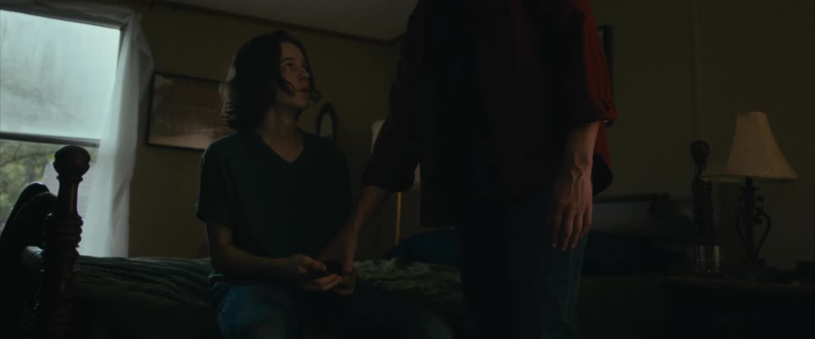 A young man sitting on a bed holding a gun looks up at a man standing next to him who takes the gun from him