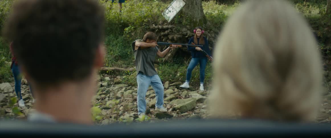A man stands in a forest and raises a shotgun and shoots as others watch and a woman claps