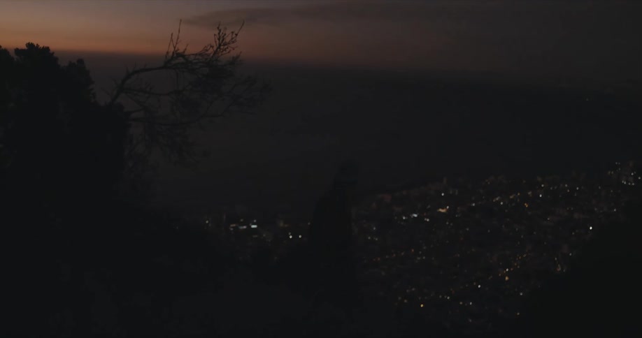 A woman at the top of a hill with a lit up city below her climbs down the hill