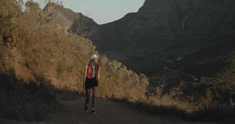 A woman wearing shorts and a sunhat is walking on a trail and there are mountains in the distance