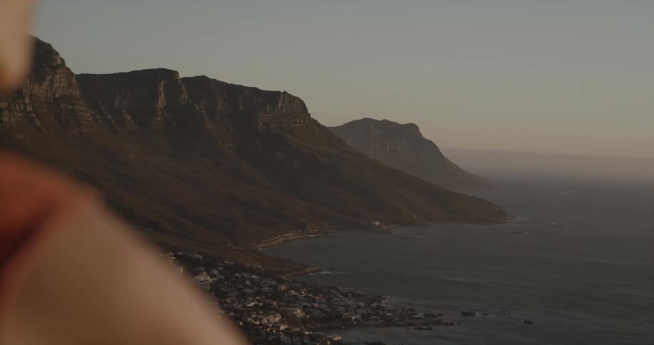 Blonde woman standing on cliff watches coastal city at dusk