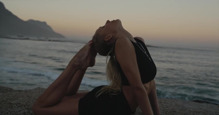 Young woman doing a yoga pose stretching her legs at the sand on the beach next to the sea