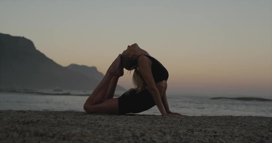 A woman is on the beach doing yoga exercises and touching her feet to her head