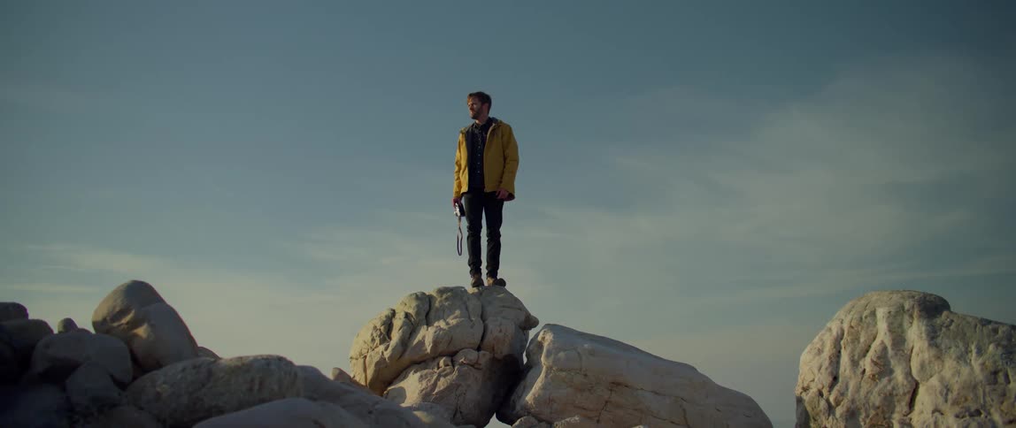 Male photographer stands on large boulder looking around before jumping down