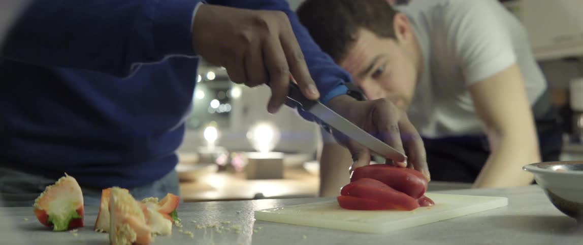 A man dices a red bell pepper while another rummages through a kitchen cabinet