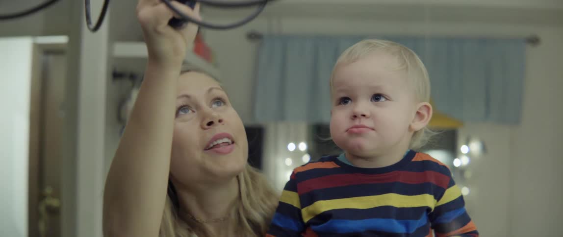 A woman holding a toddler uses a lighter on a lamp above her head