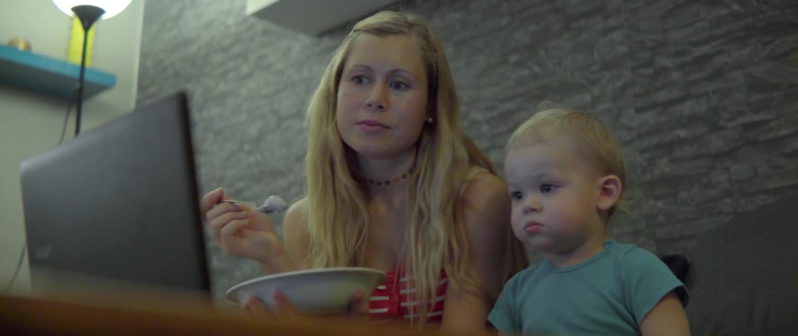 Young mother and small boy eating from a bowl while looking at a computer.