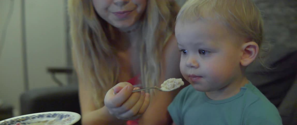 A young toddler refuses a bite of food while distracted by a television screen.