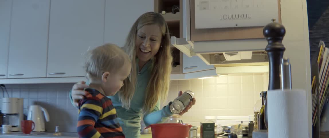 A mother and young toddler pour ingredients from a jar into a mixing bowl.