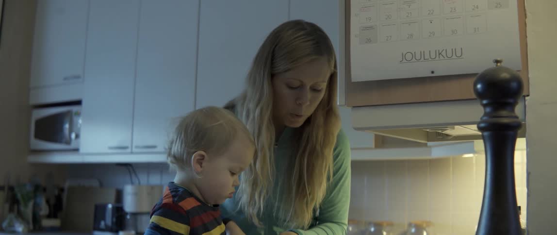 A woman pours ingredients into a red bowl while a curious toddler watches closely.