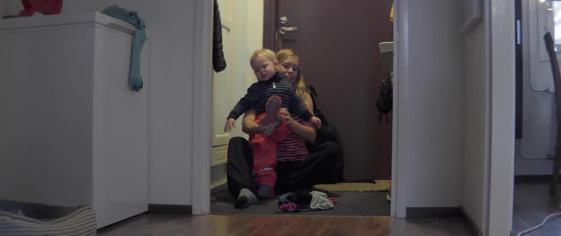 A woman sits on the floor helping her young toddler put on winter clothes.