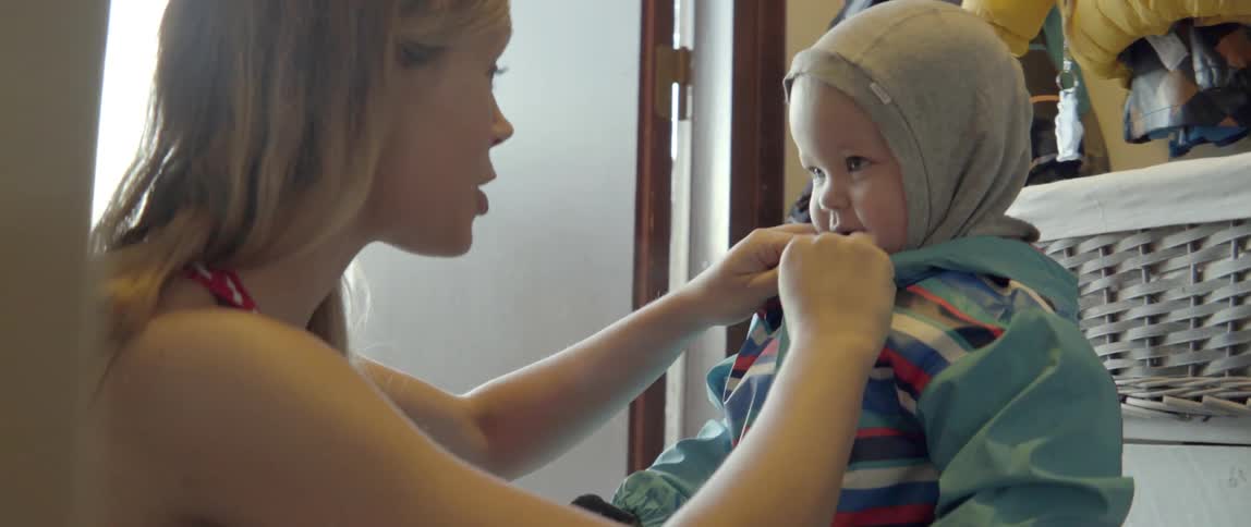 Young woman helping a small child prepare for a walk by putting on a jacket.