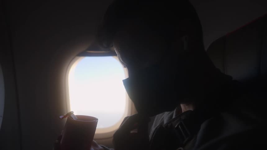 A man pulls down his mask and drinks from a cup while traveling on an airplane during the day