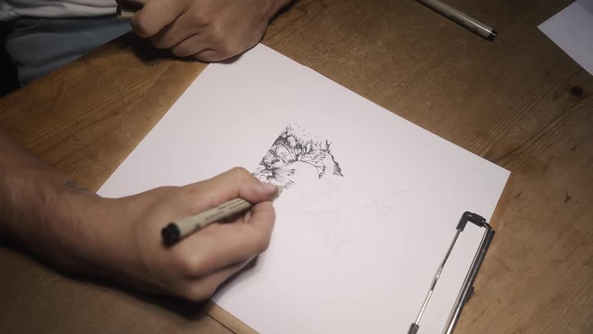 Male artist draws in ink on paper attached to clipboard set on wooden table