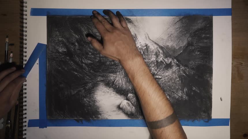 A persons left hand is peeling blue tape away from the side of a pencil or charcoal drawing that has been outlined by tape and is being held in place by the persons right arm
