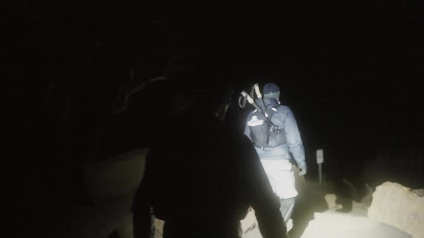 Two men are walking on a trail in the dark using a headlamp