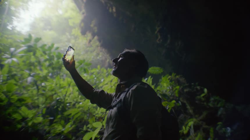 A man is taking pictures with his cell phone under a tree canopy in a jungle