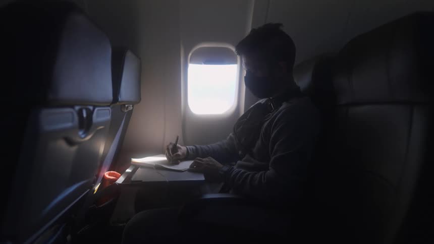 A man wearing a face mask is sitting in a seat on an airplane next to a window and writing in a book