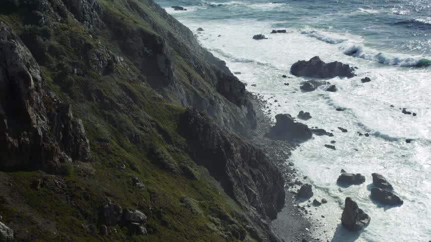 A rock strewn beach at the bottom of a mountain slope during the day