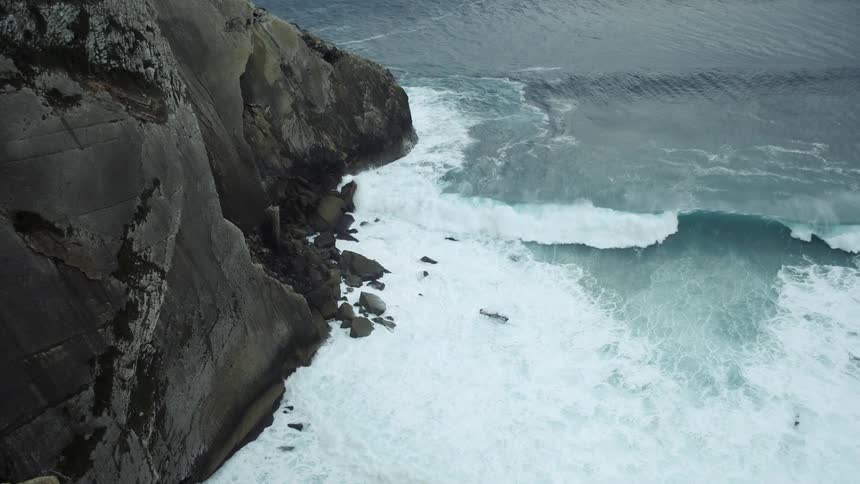View looking down from a cliff as waves roll into shore and crash on rocks along the coast
