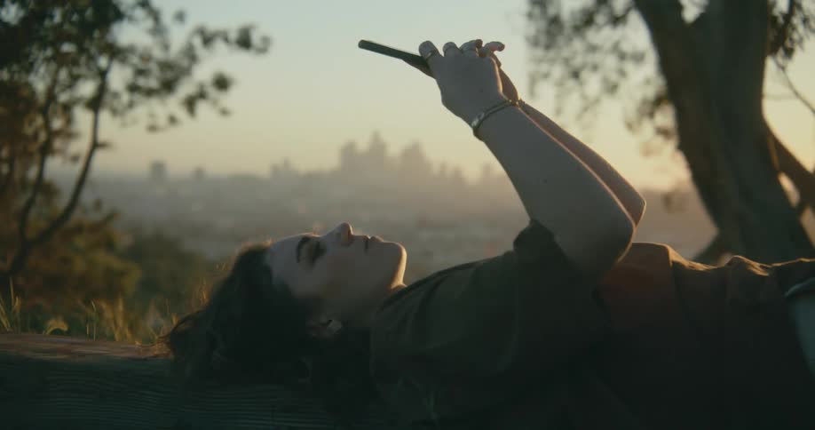 A woman looks at her cell phone as she lays on the ground near trees