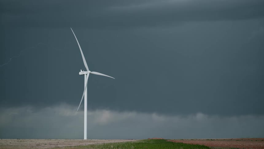 Lightning streaks through the dark clouds looming of a grassland containing a wind turbine during the day